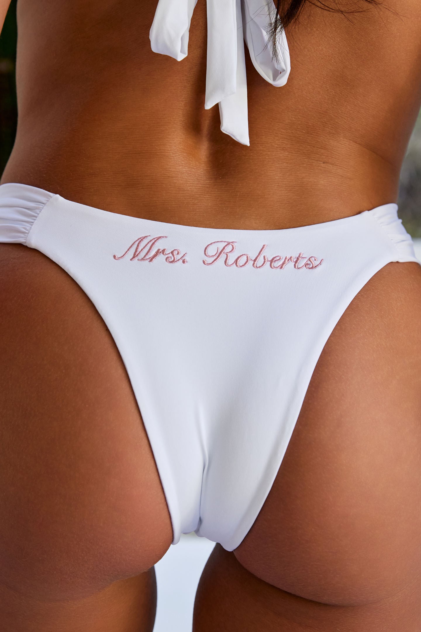Woman wearing ALT’s Mira top and Adelina bottom, a white bridal bikini set, with cacti in the background. ruched detailing. Pink embroidery Mrs. Roberts of the back of bottoms