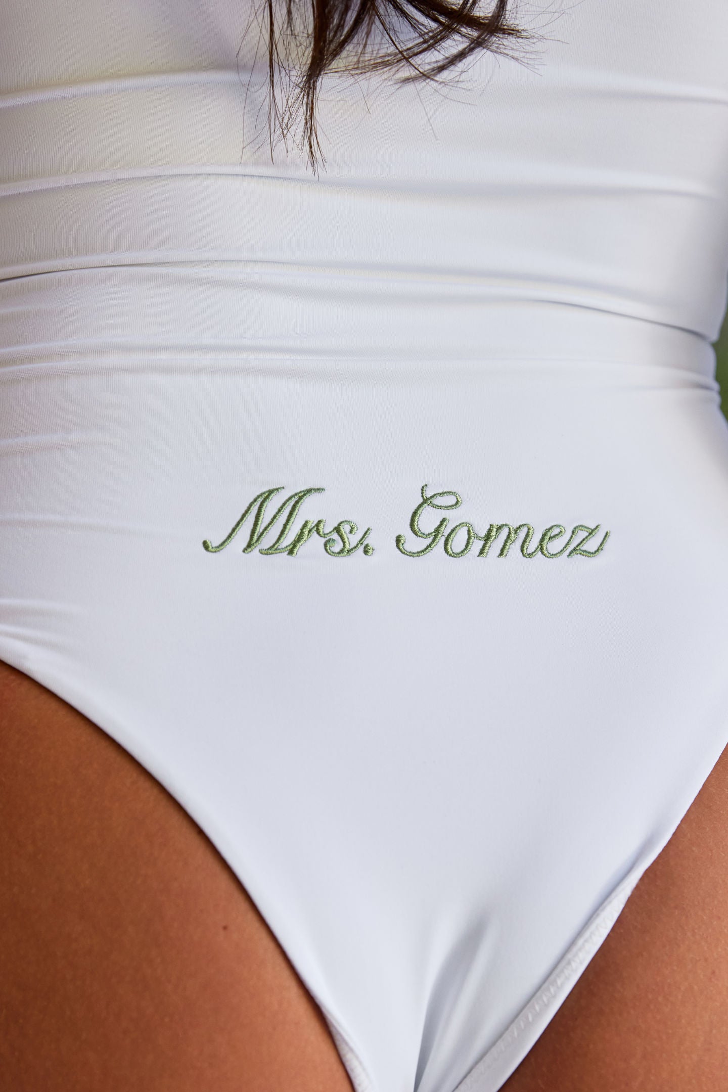 Woman wearing ALT’s Soraya one piece, a white bridal one piece swimsuit, with cacti in the background. ruching and Satin flower detailing Sage green embroidery on the back Mrs. Gomez