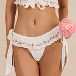 A close-up of ALT's white Mimi brief's floral ruffle trim, a personalized date on the tie, paired with a flower.