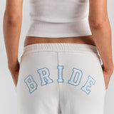 A close-up of BRIDE in heart-print varsity applique with a blue stitched outline, arched across the back of the white Bride Paola trackpant.