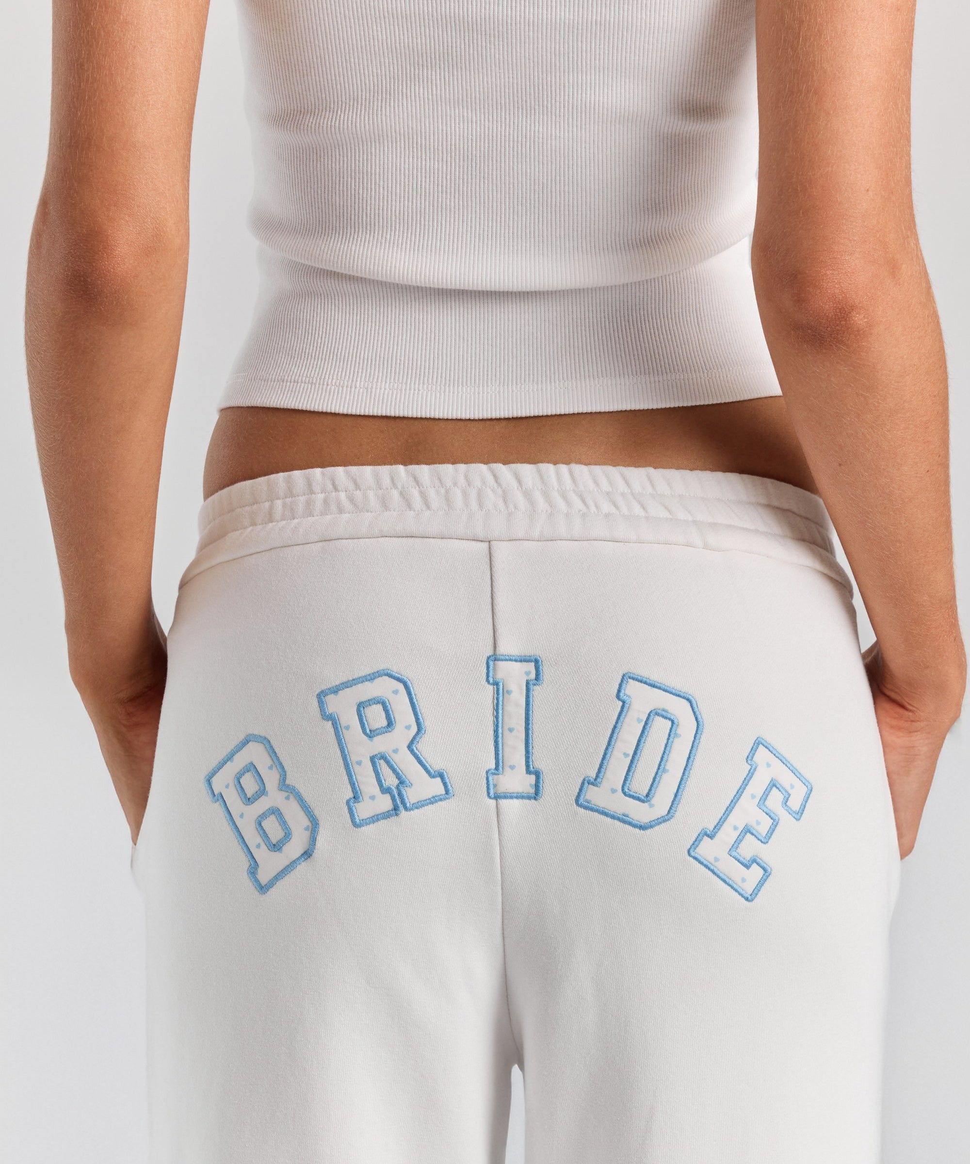 A close-up of BRIDE in heart-print varsity applique with a blue stitched outline, arched across the back of the white Bride Paola trackpant.