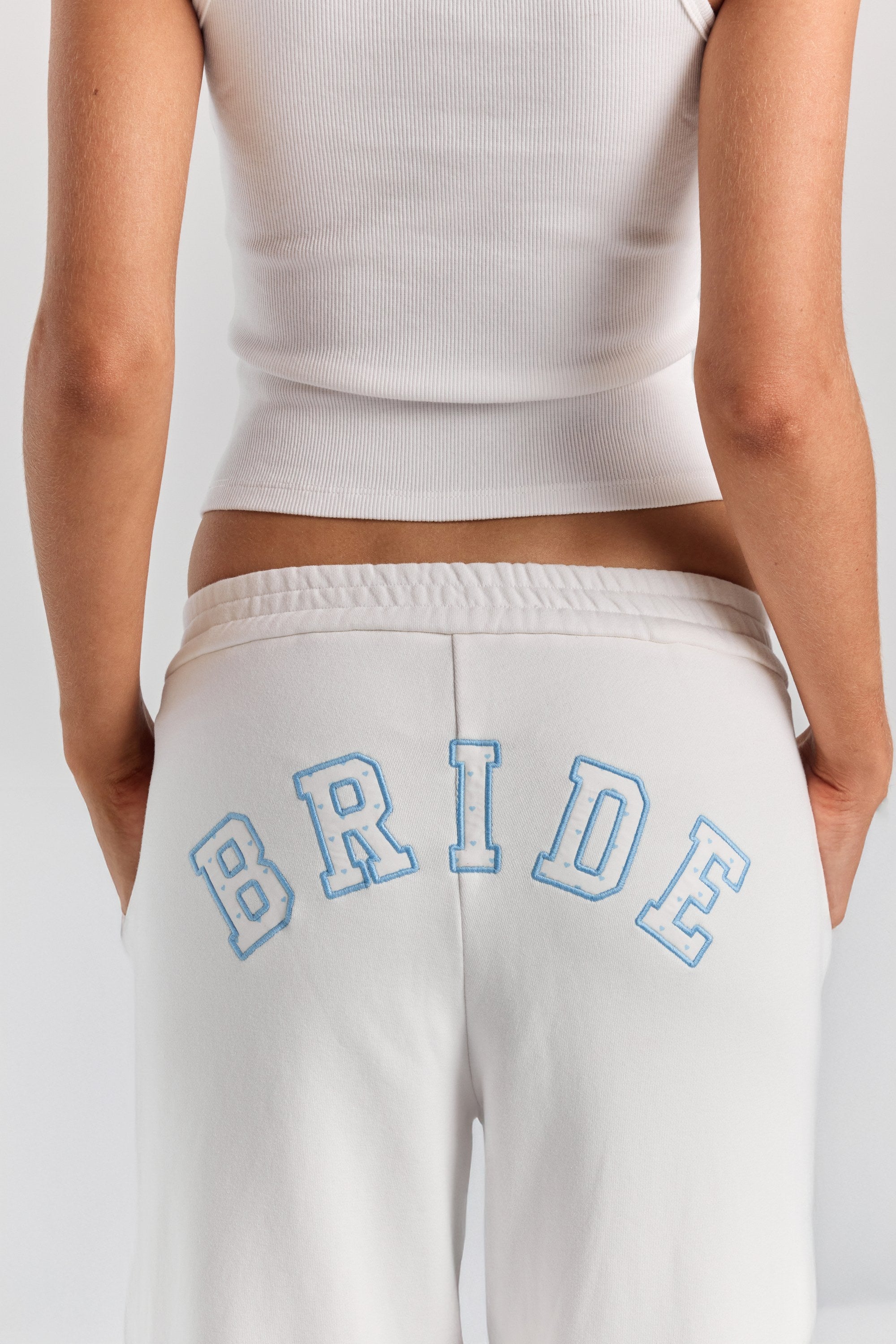 A close-up of BRIDE in heart-print varsity applique with a blue stitched outline, arched across the back of the white Bride Paola trackpant.