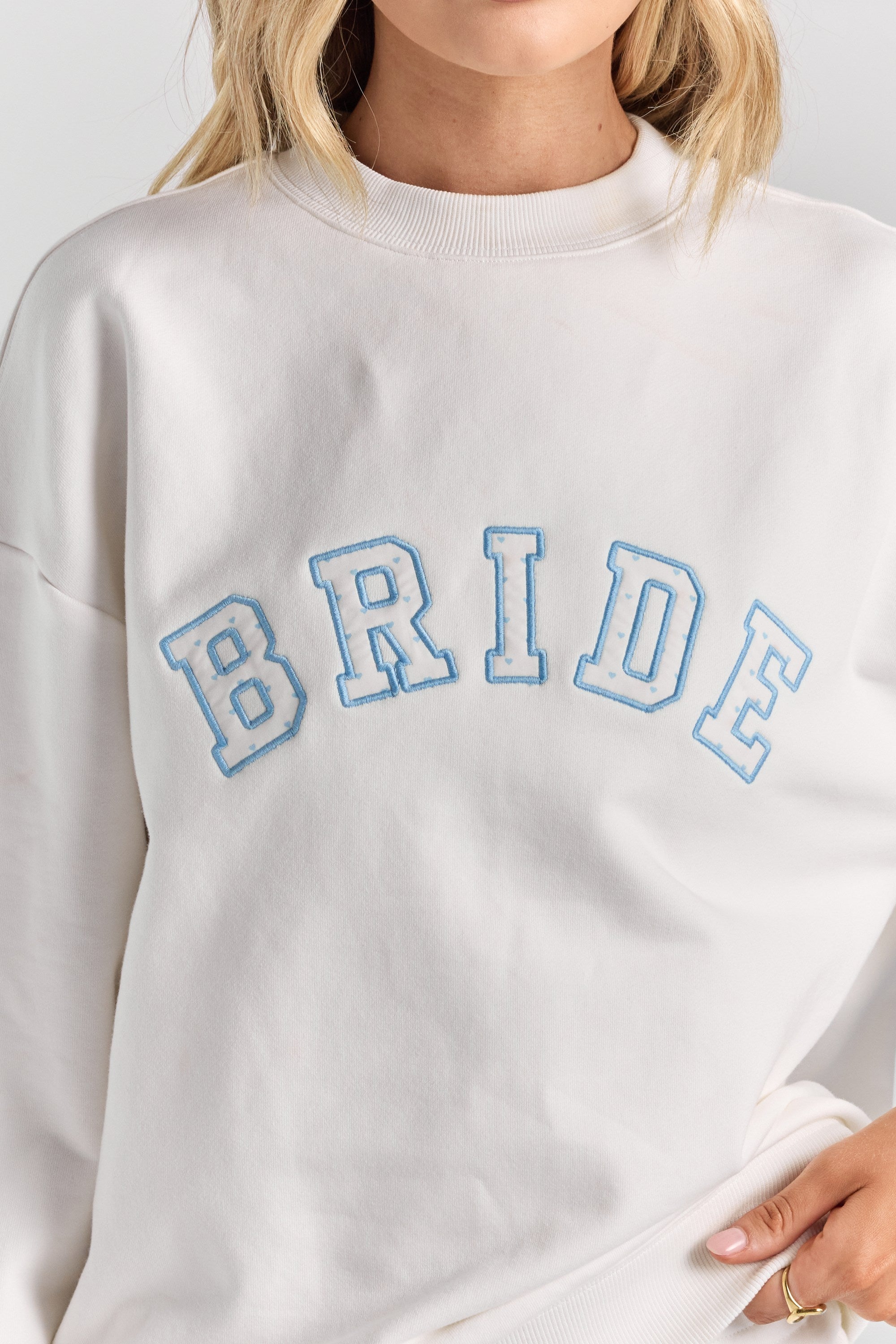A close-up of BRIDE in heart-print varsity applique with a blue stitched outline across the chest of the white Bride Pia crew sweatshirt.