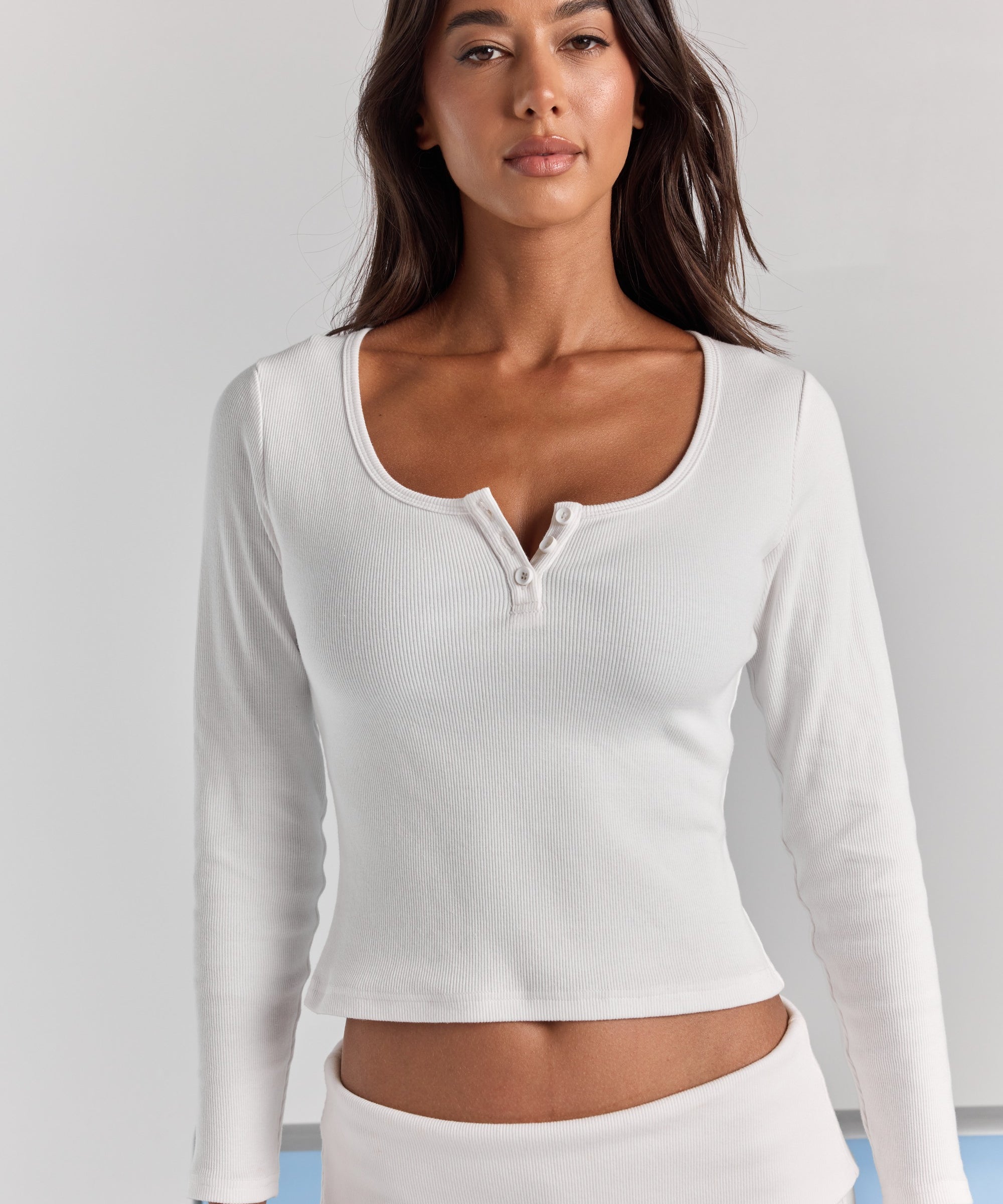 Closer front view of the white Cleo long sleeve top, with a wide scoop neck, short button placket and fitted ribbed sleeves.