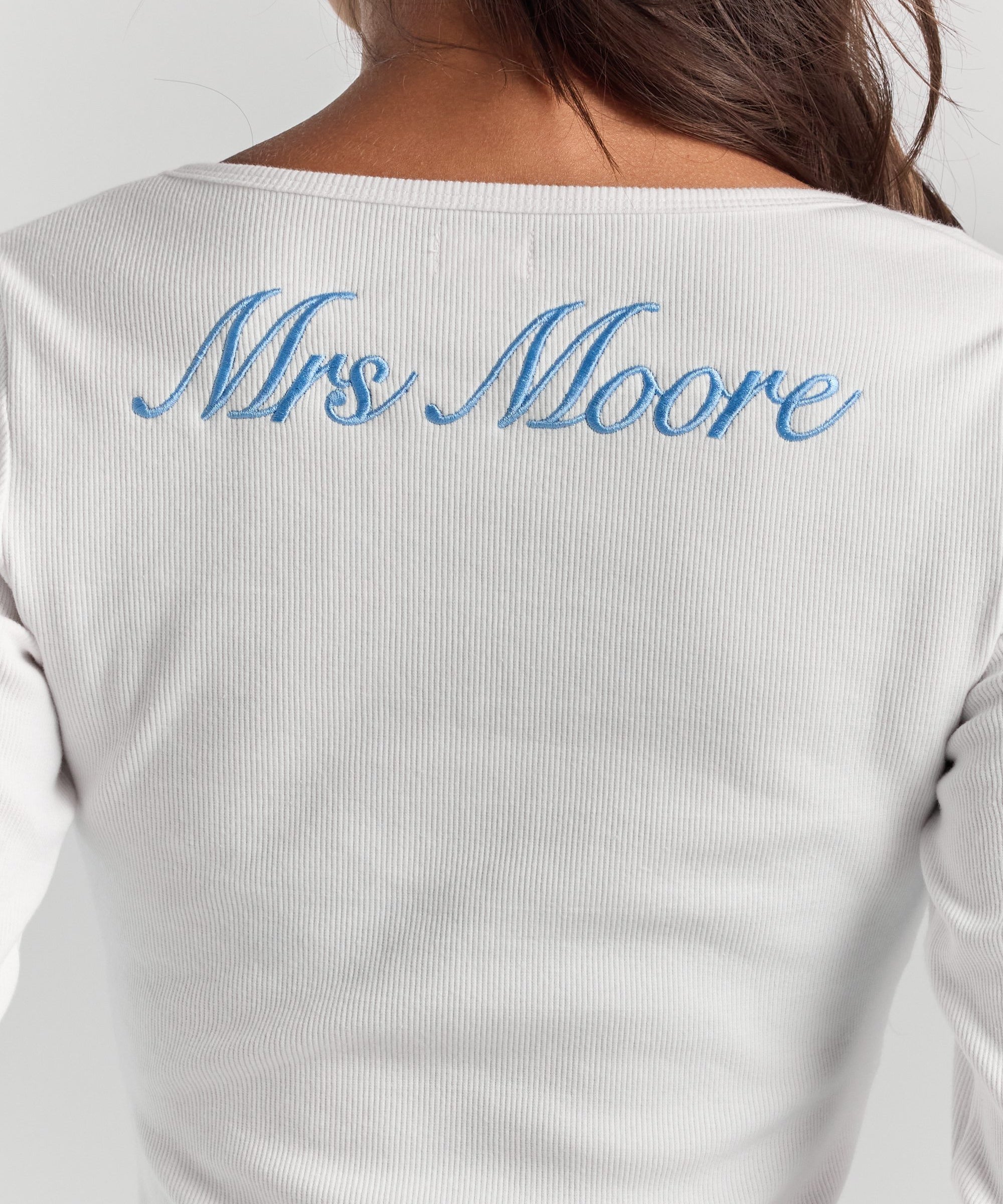 A close-up of personalized embroidery across the upper back of the white Cleo long sleeve top, just below the neckline.