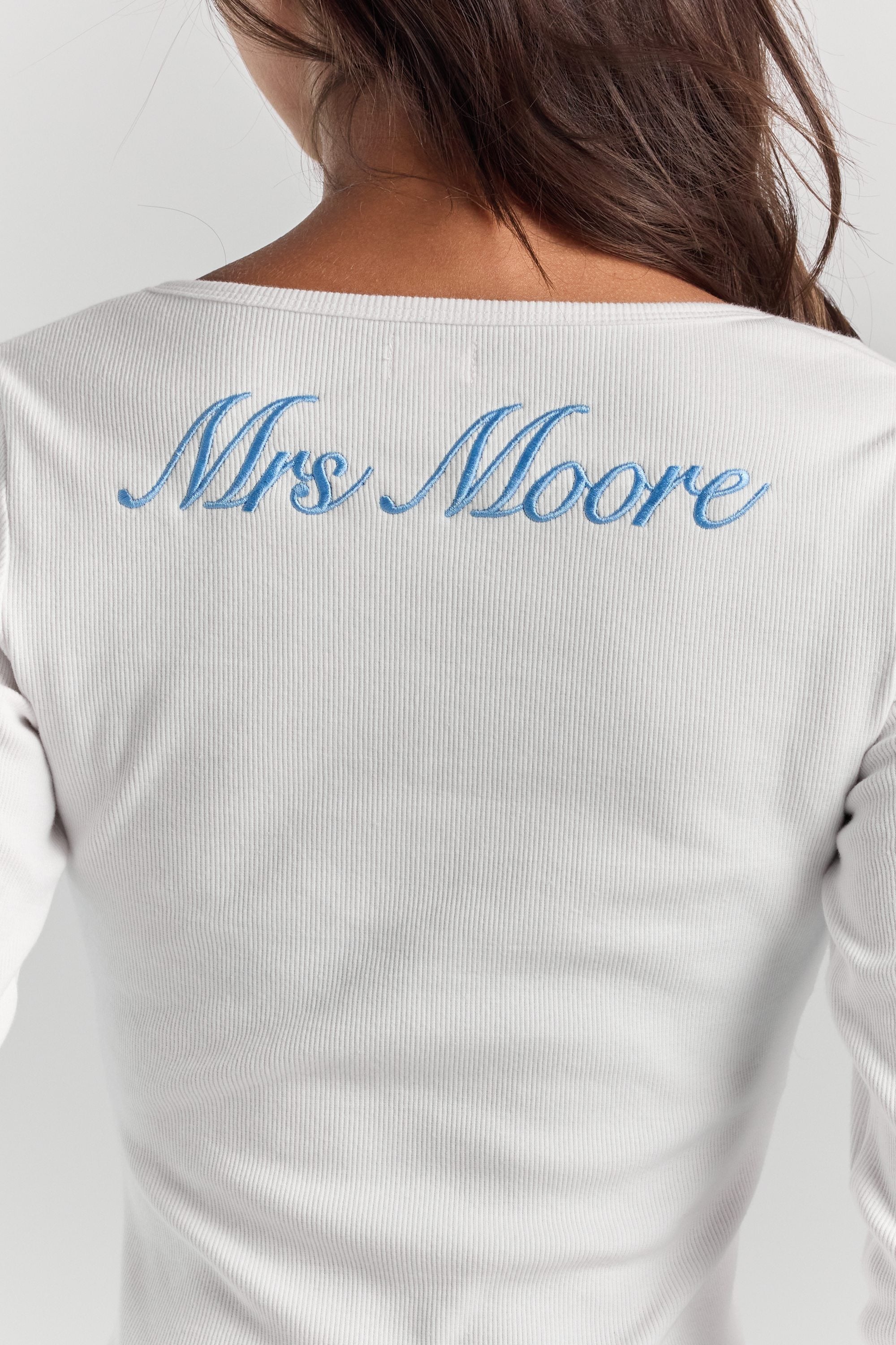 A close-up of personalized embroidery across the upper back of the white Cleo long sleeve top, just below the neckline.