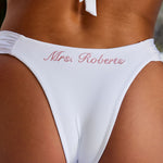 Woman wearing ALT’s Mira top and Adelina bottom, a white bridal bikini set, with cacti in the background. ruched detailing. Pink embroidery Mrs. Roberts of the back of bottoms