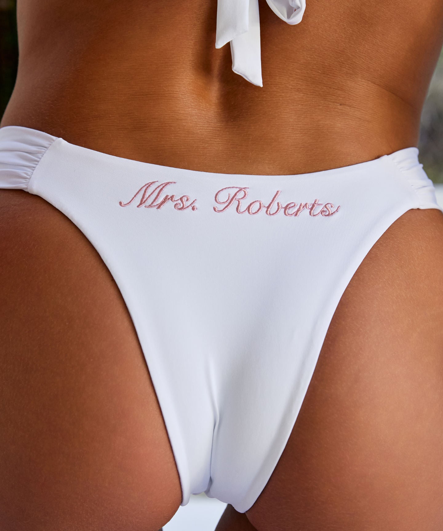 Woman wearing ALT’s Mira top and Adelina bottom, a white bridal bikini set, with cacti in the background. ruched detailing. Pink embroidery Mrs. Roberts of the back of bottoms