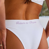 Woman wearing ALT’s Lovisa top and Esra bottom, a white bridal bikini set, with cacti in the background. Tulle flower detailing. Purple embroidery Return to travis of the back of bottoms