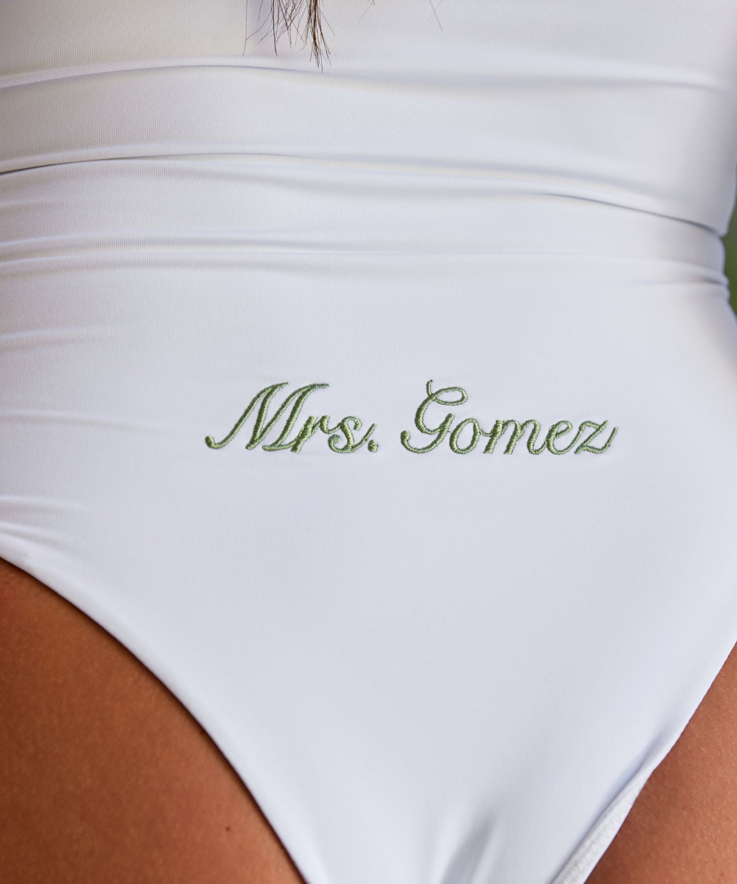 Woman wearing ALT’s Soraya one piece, a white bridal one piece swimsuit, with cacti in the background. ruching and Satin flower detailing Sage green embroidery on the back Mrs. Gomez
