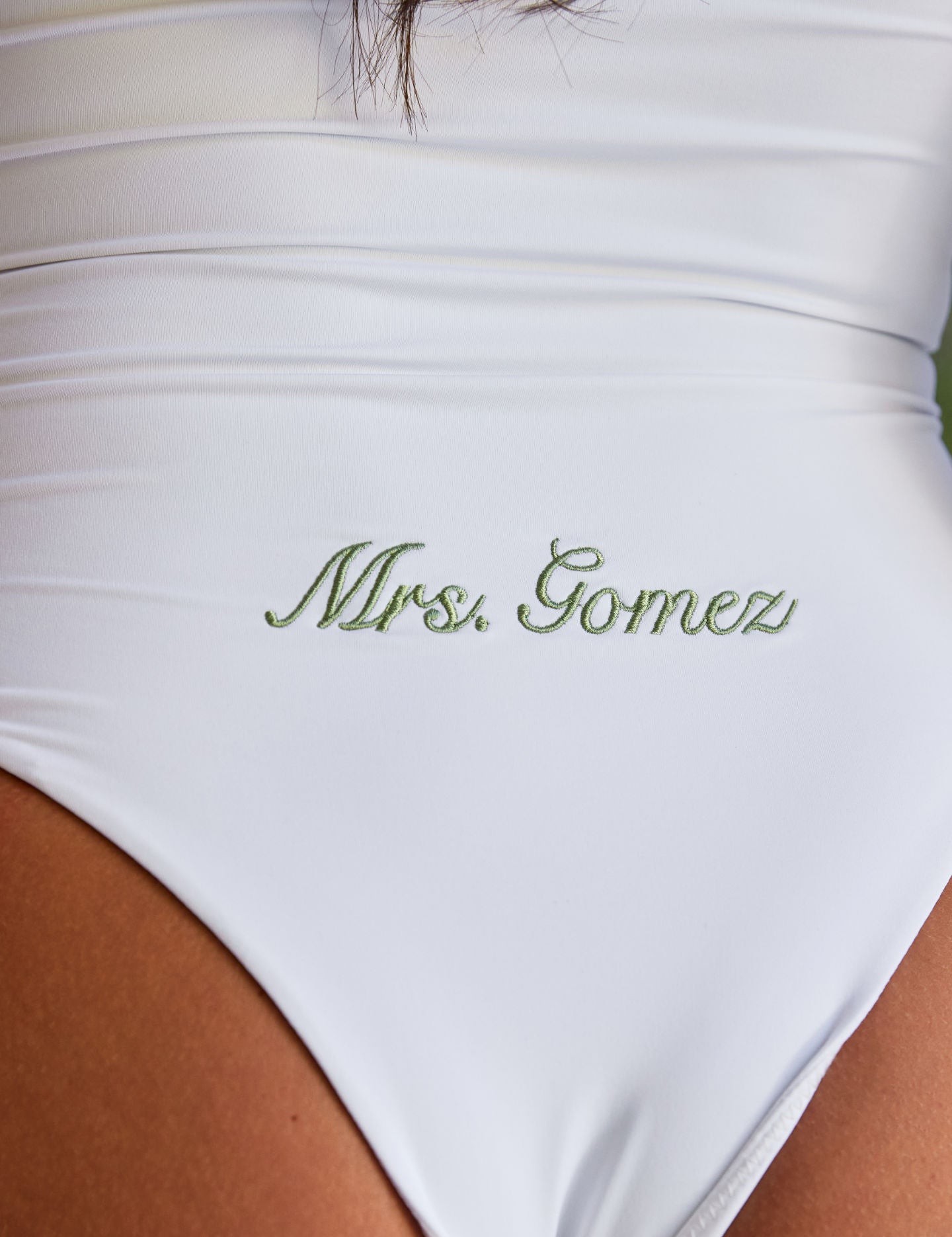 Woman wearing ALT’s Soraya one piece, a white bridal one piece swimsuit, with cacti in the background. ruching and Satin flower detailing Sage green embroidery on the back Mrs. Gomez