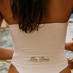 Woman wearing ALT’s Soraya one piece, a white bridal one piece swimsuit, on St. Tropez Beach. ruching and Satin flower detailing Sage green embroidery on the back Mrs. Perez