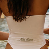 Woman wearing ALT’s Soraya one piece, a white bridal one piece swimsuit, on St. Tropez Beach. ruching and Satin flower detailing Sage green embroidery on the back Mrs. Perez