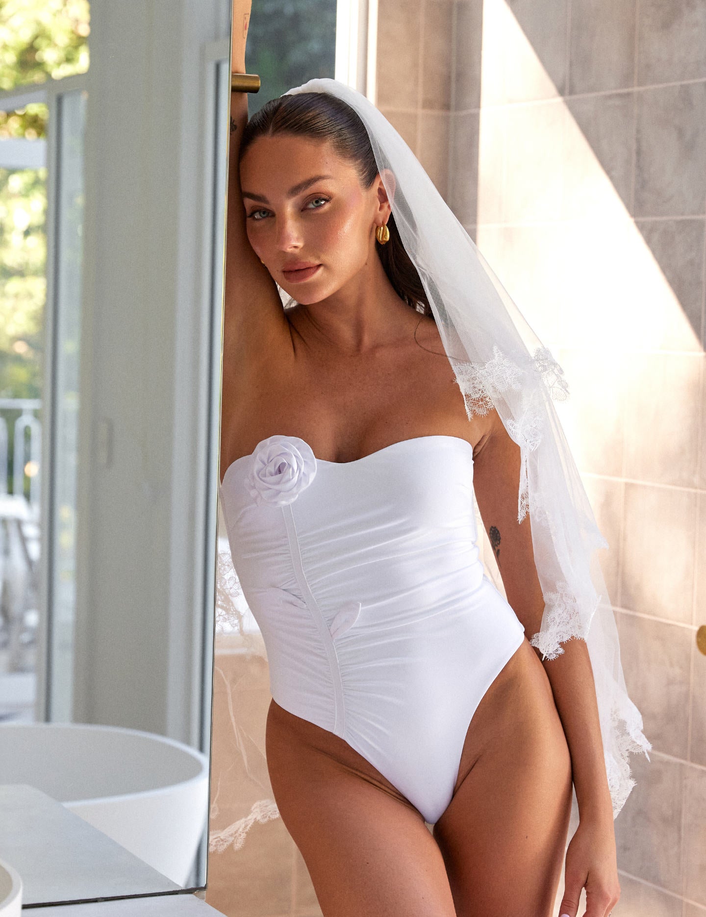 Woman wearing ALT’s Soraya one piece and florence veil, a white bridal one piece swimsuit, in luxury villa bathroom. ruching and Satin flower detailing