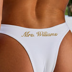 Woman wearing ALT’s Noa top and Talia bottom, a white bridal bikini set, with cacti in the background. Gold and pearl hardware. Gold embroidery Mrs. Williams on the back of bottoms